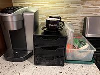 Photo showing Keurig coffee maker alongside 3-drawer pod storage and clear plastic tote containing filters and descaler, with two coffee cups with saucers on top of the drawer unit.