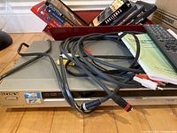 Front view of silver Sony DVD/CD player with connected AV cables, remote control, and manual booklet on top, with box of VHS tapes behind.