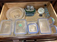 Top-down view of a drawer full of various sizes of plastic food storage containers with blue rim locking lids arranged side by side.