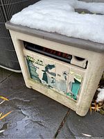 Beige Yardworks hose reel container with partially torn label and snow on top, outdoor concrete surface with leaves and nearby metal structure visible.