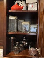 Upper shelf shows an orange polka dot teapot and white kitchen scale. Middle shelf has four clear glass canisters with white lids of varying sizes. Lower shelf contains metal lid canisters and a black and clear cookie jar labeled 'cookies'.