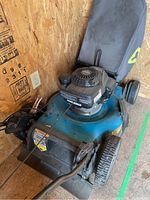 Front-right angle showing blue deck, engine housing, handlebars and attached fabric grass collector bag