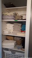 Shelves with folded linens, towels, some sheets in muted and neutral colors plus a white laundry basket at bottom.