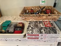 Photo showcasing woven wood basket holding eagle figures and two unopened Monopoly board games on the countertop with model trucks and cars arranged beside them.