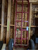 Full view of red metal hand truck with tall frame against wooden wall, showing overall structure and condition including scratches.