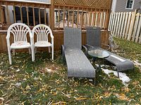 Full view of all items: two grey fabric lounge chairs, two white plastic chairs, and one glass top metal side table on grass near wooden railing with fallen leaves and snow patches.