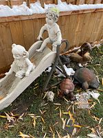 Full view of the slide featuring boy and girl statues, with several animal statues arranged around it on the grass and snow-dusted ground by a wooden fence.
