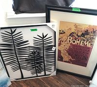 Two artworks shown side by side on the floor near a window: one black metal botanical artwork and one framed vintage styled 'La Boheme' print.
