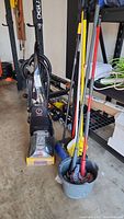 Black Bissell Ready Heat Floor Cleaner and multiple cleaning tools (mops, brooms, bucket) grouped together in a garage setting.