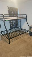 Black metal bunk bed frame with twin top bunk mattress and empty queen bottom bunk, shown from a side angle with wall and curtain background.