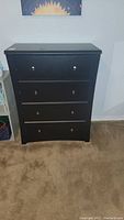 Front view of black four drawer chest of drawers against a light-colored wall on carpet.