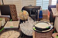Photo of large round wall clock in dark wood frame, two lidded wooden boxes with patterned carvings, white ceramic bowl with wave-like design, wooden bowls, and desk supply holders on a table in front of window blinds.