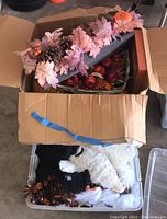 Box containing autumn leaf garlands with pinecones, artificial apples and berries; plush black and white ghost figures with orange accents, and assorted Halloween decorations.