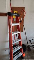 Full view of 6-foot orange Werner fiberglass ladder along with various garden and hand tools organized against the wall.