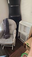 Photo of beige glider rocker chair with cushions and purple throw blanket beside white wall mount shelf and black corner shelf