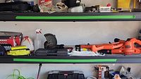 Shelf with three orange Black & Decker electric lawn tools: hedge trimmer, weed eater, leaf blower. Additional small tools and accessories nearby.
