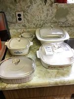 Seven Corningware baking dishes on countertop, various sizes and shapes, some with glass lids, including pieces with classic blue cornflower pattern.