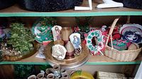 Wide view of shelf with ceramic Christmas tree, nativity plaques, handmade wreath, coasters, Santa figurine, chargers, paper plates, and tinsel garlands.