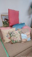 Overview of eight decorative pillows of various styles and colors arranged on a chair including embroidered horse image, solid colors, floral cross stitch, vintage knit textures.