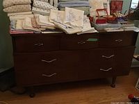 Front view of the wood dresser with seven drawers showing the brown finish and metallic handles, top surface holding linens and other items.