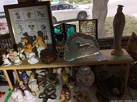 Full view of wooden shelves showing various Goebel Hummel porcelain figurines, Zabkowice glass horse statue, small ceramic vases, and other decorative objects in natural daylight from a window.
