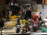 Full view of kitchenware lot showing utensils, containers, measuring cups, placemats, and various kitchen tools piled on a countertop.