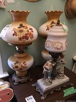 Photo showing two beige glass faux oil lamps with sunflower and leaf painted designs, scalloped tops, and ornate metal bases next to a figurine lamp with a white and gold glass shade on a stone base with porcelain figurines of a man and woman.