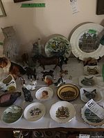 Overview of lot on table showing various Beswick horse figurines, owl and collie animal figurines, commemorative and scenic decorative plates, including Chadwick cat head plates