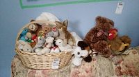 Basket filled with assorted small plush toys including bears and dogs, some with heart tags inside a wicker basket, plus a large brown teddy bear and Steiff chicken outside the basket on a floral couch