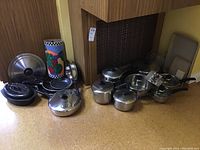 Overview of cookware assortment including stainless steel saucepans, nonstick frying pans, enamel roaster, sauté pan, and decorative container