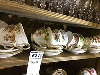 Shelf displaying a variety of porcelain teacups and saucers with floral and gold decorative patterns, stacked neatly in pairs.