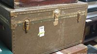 Side view of vintage trunk showing metal trim and latches with a label attached to front.