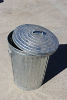 Large galvanized metal bin with circular ridged lid partially open showing interior, placed on concrete surface.