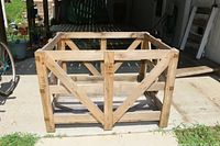 Open rectangular wooden pallet crate box on concrete surface outside. Natural wood slats with cross-bracing and reinforced corners.