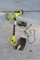 Overall view of Ryobi cordless string trimmer/edger with attached battery, charger and power cord laid on concrete surface.