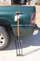Full view of the antique curd mixer leaned against a green truck, showing wooden handle with attached tag and large metal paddle with three slots.
