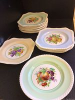 Overview showing four stacks of square plates in blue, green, pink colors and one green round plate, all with fruit motifs and rope edge design.