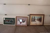 Three framed artworks arranged on the floor showing the C.M. Bowman painting on right, needlework in middle, and Steve Childs print on left.