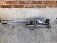 Top view showing the full length metal handle with black grip and steel body of the trolley jack resting on concrete ground against a brick wall.