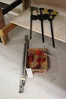 Four metal pipe clamps (two 26 inch pipes, two 33.5 inch pipes) with red clamp heads in a box, on a concrete floor near wooden furniture.