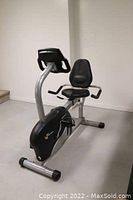Side view of the recumbent exercise bike showing seat with backrest and armrests, pedals and frame structure.