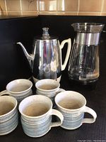 Photo showing GE Electric percolator with white handle, Pyrex Corning glass carafe with metal trim, and six pottery mugs in speckled beige and blue color.