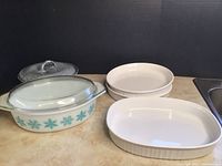 Photo of vintage Pyrex Snowflake oval casserole dish with clear lid, and white Corningware oval and round baking dishes on countertop.