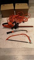 Electric Black & Decker hedge trimmer, orange with black handle and dual blades, laid flat with extension cord behind it and two hand saws below.