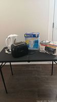 Photo showing white electric kettle, black toaster, Brita water filter pitcher box, and Chef's Choice electric knife sharpener box on black table