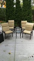 4 patio chairs with striped reversible cushions and 2 small round side tables with glass tops arranged on concrete patio outdoors with greenery background.