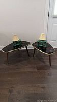 Two mid-century modern style side tables with black glass tops featuring gold and red swirling patterns, each with three legs and two green glass lamps with beige lampshades placed on top.