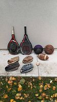 Full lot showing tennis rackets, basketballs, football, baseball gloves and baseball on white surface outdoors with leaves on ground.