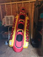 Front view of deflated inflatable towable tube standing upright against plywood wall, showing overall shape and graphics.