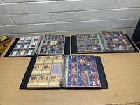 Three black binders open on a table showing multiple pages of assorted basketball cards in plastic sleeves, various players and teams visible.
